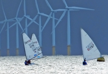 The lead boats run out to sea under a leaden sky, in the shadow of the windfarm